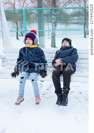 Two children bundled in winter wear sit on a snowy swing, enjoying a winter day outdoors. 127442829