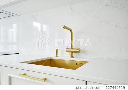 Stylish modern kitchen sink with gold faucet and white marble countertop during morning light Stylish modern kitchen sink with gold faucet and white marble countertop during morning light 127444319