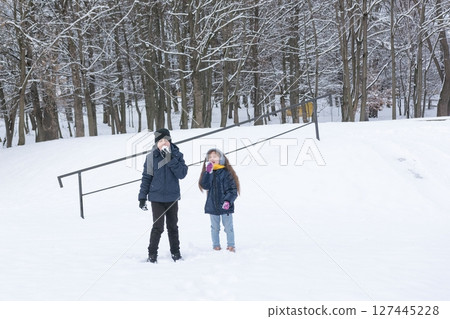 Two children enjoy a snowy day, playfully eating snow in a winter park setting. Two children enjoy a snowy day, playfully eating snow in a winter park setting. 127445228