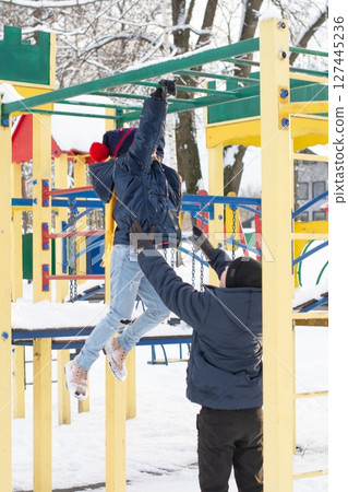 A child hangs from a playground bar while an adult assists, enjoying a snowy winter day. A child hangs from a playground bar while an adult assists, enjoying a snowy winter day. 127445236