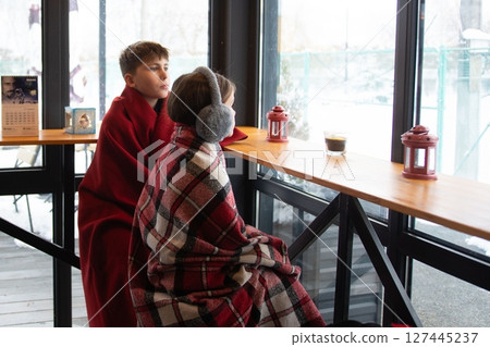 Two children snuggled in blankets watch the snowy landscape from a warm window seat. Two children snuggled in blankets watch the snowy landscape from a warm window seat. 127445237