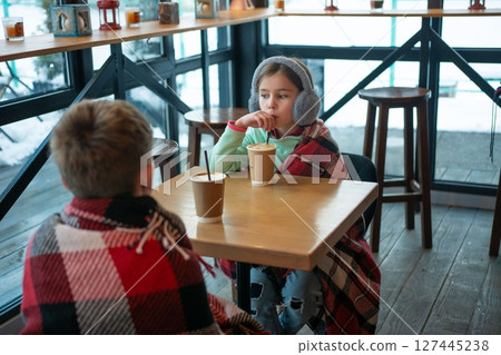 Two children enjoy warm drinks at a cafe, wrapped in cozy blankets near a window overlooking a snowy scene. Two children enjoy warm drinks at a cafe, wrapped in cozy blankets near a window overlooking a snowy scene. 127445238