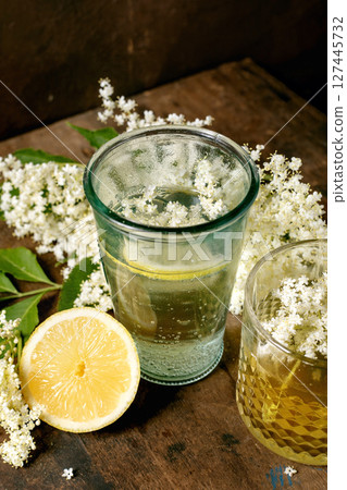 Refreshing elderflower beverage in glass with fresh lemon slice and white elderflower blossoms on wooden background. Natural herbal drink concept. 127445732