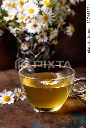 Glass cup chamomile tea decorated daisy flower fresh flowers vase wooden table background. Herbal beverage natural wellness concept. Glass cup chamomile tea decorated daisy flower fresh flowers vase wooden table background. Herbal beverage natural wellness concept. 127445734
