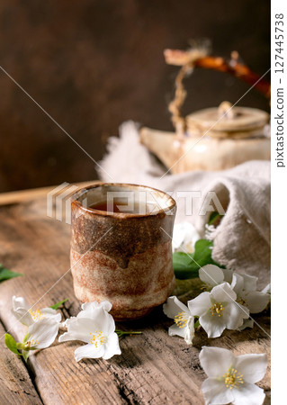 Ceramic tea cup with hot tea white spring flowers teapot on wooden table. Traditional oriental tea ceremony setup with natural decoration. Ceramic tea cup with hot tea white spring flowers teapot on wooden table. Traditional oriental tea ceremony setup with natural decoration. 127445738