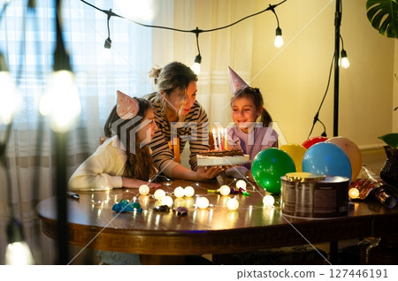 Celebration of joy and laughter as a woman presents a birthday cake to two delighted children surrounded by colorful decorations at a cozy indoor gathering in the evening 127446191