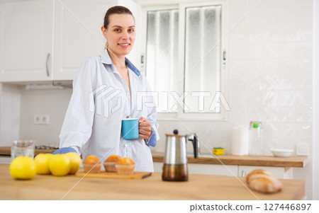 Smiling young woman in male shirt drinking coffee in home kitchen 127446897