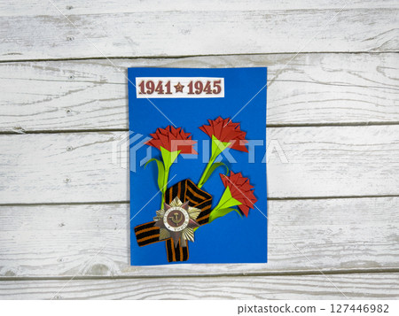Handmade Victory Day card with red paper carnations, St. George ribbon, and WWII medal on blue background. The years 1941-1945 symbolize the Great Patriotic War. Craft tribute on white wood Handmade Victory Day card with red paper carnations, St. George ribbon, and WWII medal on blue background. The years 1941-1945 symbolize the Great Patriotic War. Craft tribute on white wood 127446982