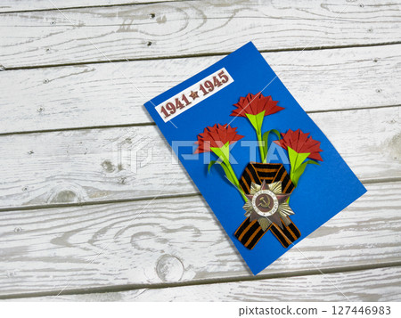 Victory Day greeting card with red paper carnations, St. George ribbon, and Soviet WWII medal on blue background. 1941 - 1945 date emphasizes memory of the war. Handmade craft on wooden surface 127446983