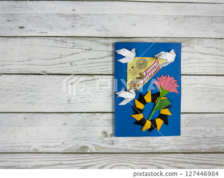 Handmade Victory Day card with origami doves, pink carnation, laurel wreath, and I remember Im proud banner on blue background. Symbol of peace and memory on rustic wooden surface Handmade Victory Day card with origami doves, pink carnation, laurel wreath, and I remember Im proud banner on blue background. Symbol of peace and memory on rustic wooden surface 127446984