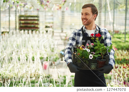 Black uniform, holding flowers. Man is working in garden center. Successful employee is in a bright greenhouse Black uniform, holding flowers. Man is working in garden center. Successful employee is in a bright greenhouse 127447078
