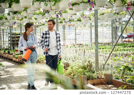 Walking, looking at each other, holding watering device. Florist man and woman are together in greenhouse 127447278