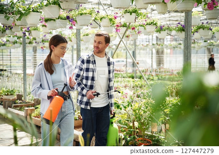 Walking, looking at each other, holding watering device. Florist man and woman are together in greenhouse 127447279