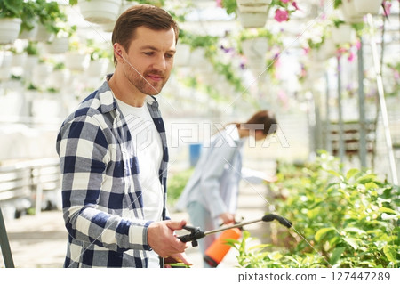 Holding watering cans. Florist man and woman are working together in bright greenhouse 127447289