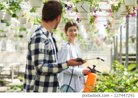 Holding watering cans. Florist man and woman are working together in bright greenhouse 127447291