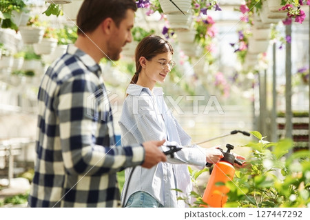 Holding watering cans. Florist man and woman are working together in bright greenhouse 127447292