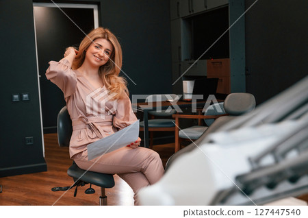 Sitting and holding document. Beautiful young female doctor is indoors in the cabinet Sitting and holding document. Beautiful young female doctor is indoors in the cabinet 127447540