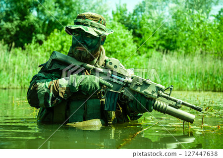 Soldier in camouflage holding a rifle partially submerged in water with green vegetation in the background under daylight. 127447638