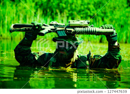 Camouflaged soldier in aquatic setting holding a rifle above head while partially submerged in water amidst lush greenery. 127447639