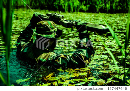 Soldier in camouflage wades through a swamp holding a rifle above the water surface surrounded by aquatic plants and greenery. 127447646