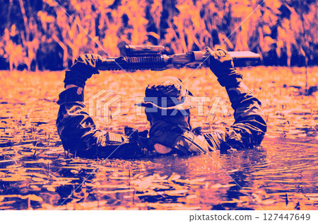 A soldier wearing camouflage holds a firearm above his head while wading through water surrounded by reeds in a dramatic color filter. 127447649