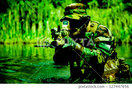 Soldier in camouflage uniform and hat aiming a rifle with a scope while wading through a green forested water area. 127447656