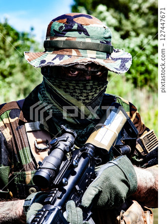 Soldier with camo gear and face covering, holding a rifle with optics in a green, wooded outdoor setting under clear blue sky. 127447671