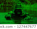 Soldier in full camouflage gear and bucket hat holding a knife, crawling through a body of water with vegetation in the background. 127447677