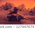 Soldier in camouflage gear and hat, holding a rifle, wading through a swamp with trees in the background under an orange-tinted sky. 127447679