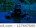 Man in camouflage clothing and hat crouching in water holding a knife with foliage in the background. 127447680