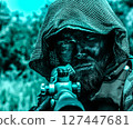 Intense sniper with a hood and camouflage face paint aiming a rifle, set against a blurred natural background with a blue color tone. 127447681