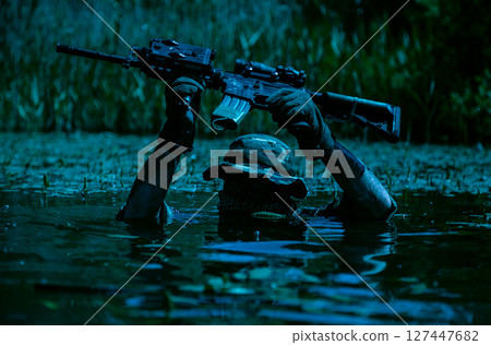 Soldier in camouflage gear and hat holding a rifle above water in a wetland area during nighttime. Vegetation is visible in the background. 127447682