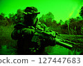 Soldier in camouflage with a rifle crouches in water surrounded by dense vegetation, wearing a hat and gloves, during low-light conditions. 127447683