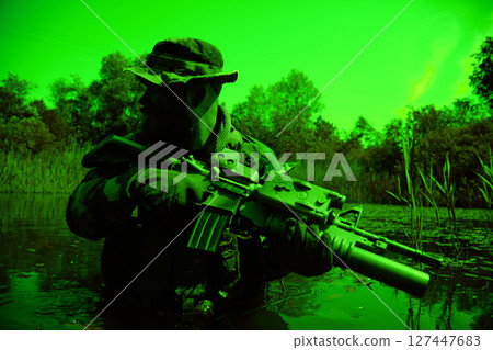 Soldier in camouflage with a rifle crouches in water surrounded by dense vegetation, wearing a hat and gloves, during low-light conditions. Soldier in camouflage with a rifle crouches in water surrounded by dense vegetation, wearing a hat and gloves, during low-light conditions. 127447683
