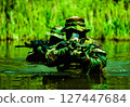 Soldiers in camouflage uniforms with rifles navigate through water in a dense green vegetation setting, demonstrating stealth and tactical movement. 127447684
