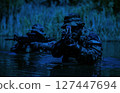 Two armed soldiers in camouflage uniforms navigate through water with rifles aimed forward, under a low-light environment and surrounded by vegetation. 127447694