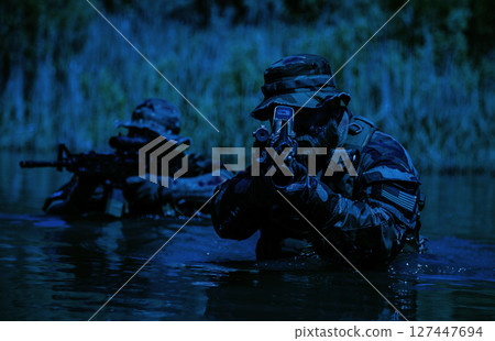 Two armed soldiers in camouflage uniforms navigate through water with rifles aimed forward, under a low-light environment and surrounded by vegetation. 127447694