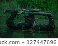 Camouflaged soldier with face paint held rifle above head while wading through water in a dense forest environment under daylight. 127447696