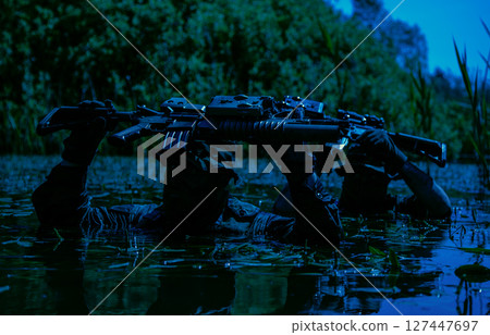 Two armed soldiers in camouflage, carrying rifles above water, wade slowly through a dense, swamp-like area surrounded by greenery. 127447697