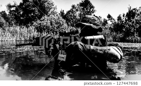 Silhouette of a soldier submerged in water, holding a rifle with foliage and trees in the background under a clear sky. 127447698