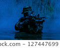 Soldier in camouflage gear wading in water, holding a rifle with tactical equipment, surrounded by dense vegetation under a blue-tinted light. 127447699