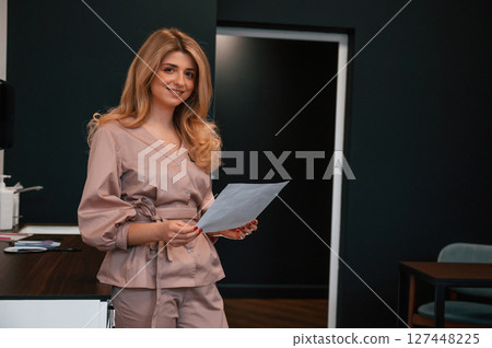 Standing and holding document. Beautiful young female doctor is indoors in the cabinet Standing and holding document. Beautiful young female doctor is indoors in the cabinet 127448225