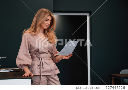 Standing and holding document. Beautiful young female doctor is indoors in the cabinet Standing and holding document. Beautiful young female doctor is indoors in the cabinet 127448226