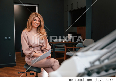 Sitting and holding document. Beautiful young female doctor is indoors in the cabinet Sitting and holding document. Beautiful young female doctor is indoors in the cabinet 127448232