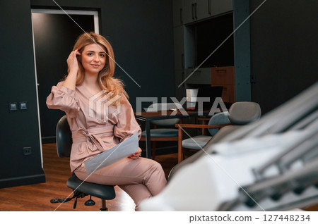 Sitting and holding document. Beautiful young female doctor is indoors in the cabinet 127448234