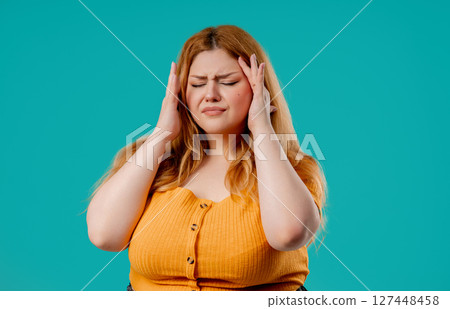 Plus size woman touching temples, suffering from headache, blue studio. Pain. 127448458