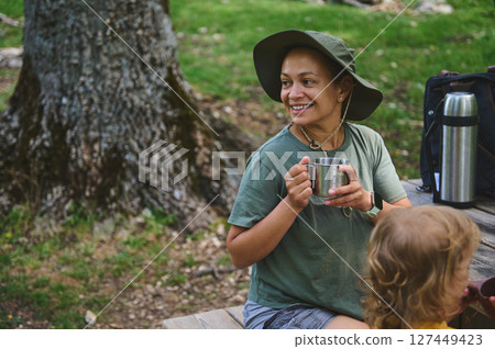 Smiling Woman Enjoying Coffee Outdoors with Child in a Scenic Park Setting 127449423