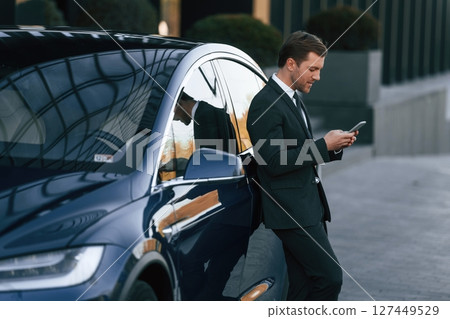 Using smartphone. Businessman is standing near his car outdoors 127449529