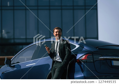 Smiling, holding smartphone. Businessman is standing near his electric car outdoors Smiling, holding smartphone. Businessman is standing near his electric car outdoors 127449535
