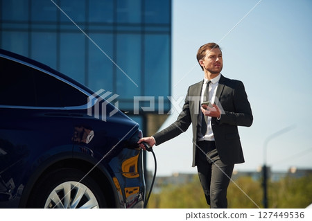 Holding the charger. Businessman is standing near his electric car outdoors Holding the charger. Businessman is standing near his electric car outdoors 127449536